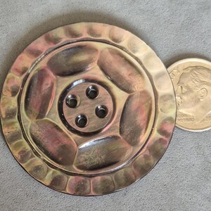 May include: A metallic, circular object with a textured rim and a central design featuring four holes. A U.S. dime is placed next to the object for size comparison. The object has a bronze and gold color.