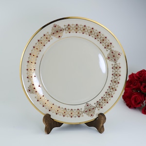 May include: A white ceramic plate with a gold rim and decorative border featuring a pattern of gold and burgundy accents. The plate is displayed on a wooden stand, with a bouquet of red roses in the background.