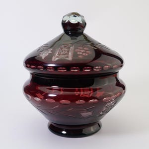 May include: A vintage glass candy jar with a lid. The jar is made of dark red glass and has a cut glass design of grapes and leaves. The lid has a small knob on top.