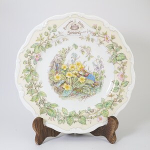 May include: A white ceramic plate with a floral design featuring a cottage, primroses, and a small rabbit. The plate has a scalloped edge and the words "Beneath a Hedge" and "Spring" are printed on the plate.