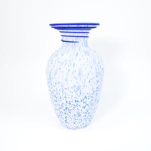May include: A glass vase featuring a white base with blue speckled patterns throughout the body. The neck of the vase is narrow with a flared rim, decorated with several horizontal blue glass rings that wrap around the top section.