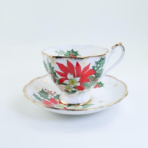 May include: A white teacup and saucer set with a festive Christmas poinsettia design. The teacup features a red poinsettia, green holly, and gold trim. The saucer has a matching design and gold edging, creating an elegant holiday aesthetic.