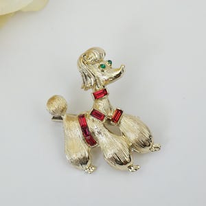 May include: Gold-tone brooch of a poodle with red and green rhinestones. The poodle is standing on all four legs with its tail curled up.