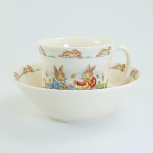 May include: Cream-coloured ceramic cup and bowl set. The cup has a handle and is decorated with illustrations of rabbits and a scene with two rabbits. The bowl has a wide, shallow design. Likely for children.