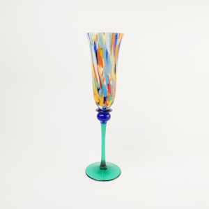 May include: A tall, slender glass flute featuring a flared rim and a bowl decorated with a multicoloured speckled pattern of orange, blue, and yellow. The stem is a solid green glass, connected to the bowl by a small, round, deep blue glass bead.