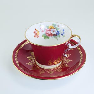 May include: A red teacup and saucer set with gold detailing and floral patterns. The teacup has a white interior with a colourful floral design. The saucer features a gold leaf pattern around the rim. A classic tea set.
