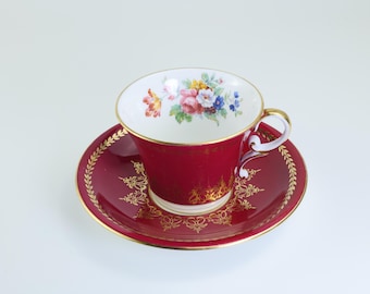 May include: A red teacup and saucer set with gold trim and floral accents. The teacup features a white interior with a colorful floral design. The saucer has a gold leaf pattern around the edge. A classic tea set.