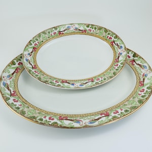 May include: Two oval serving platters. The white platters have a decorative border with a gold rim. The border features a design of birds, flowers, and foliage in shades of green, red, and blue.