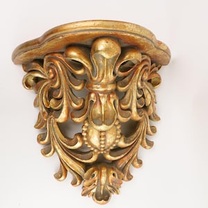 May include: A wall-mounted shelf featuring a semi-circular top surface and an ornate, carved support bracket. The piece is finished in a metallic gold tone with darker, distressed accents highlighting the intricate scrollwork and leaf motifs.
