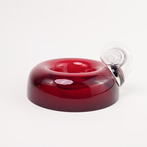 May include: A deep red, circular glass bowl with a smooth, doughnut-like shape and a central indentation. Attached to one side of the red glass body is a clear, transparent glass handle shaped like a coiled, spiralling shell or scroll.