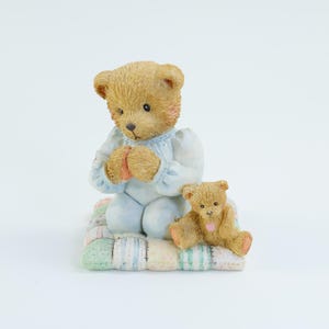 May include: A figurine of two teddy bears. One bear is kneeling in prayer, wearing a light blue outfit, and the other is sitting. They are on a pastel-colored quilt. The bears are light brown with pink accents.