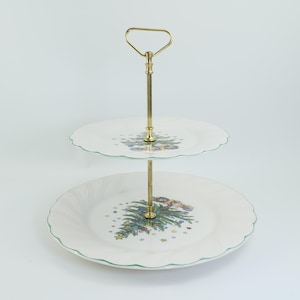 May include: Two-tiered serving stand with a gold-coloured handle and support. The white plates have a scalloped edge with a green trim and a Christmas tree design. Ideal for serving desserts or appetizers.