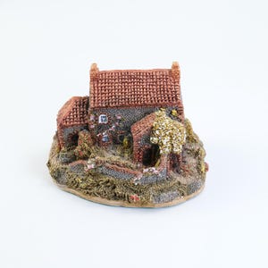 May include: A miniature house figurine with a red tile roof, stone walls, and small windows. The base is textured with greenery and a small tree. The overall color palette includes shades of brown, red, and green.