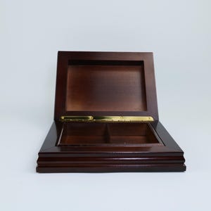 May include: A dark brown wooden box, rectangular in shape, with a hinged lid and brass-coloured hinge. The interior is divided into two compartments. The box is open and sits on a white surface.