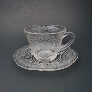 May include: A clear glass teacup and saucer set with an intricate floral pattern.