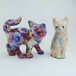 May include: Two ceramic cat ornaments. One is walking, the other is sitting. Both cats feature a floral pattern in purple, blue, and white, with green detailing. The ornaments are decorative.