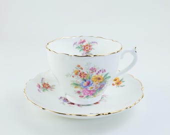 May include: White teacup and saucer set with a floral design. The cup and saucer have a scalloped edge and gold trim. The floral pattern includes pink, yellow, blue, and orange flowers.