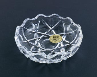 May include: A clear, decorative crystal bowl with a scalloped rim and geometric patterns. The bowl has a sticker that reads "RCR Cristallo Italiano 24% PbO". The bowl is on a dark surface.