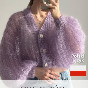 May include: A cropped, lavender-coloured, open-knit cardigan featuring long, voluminous sleeves, a V-neckline, and three iridescent buttons down the front. The garment is made of a fuzzy, textured yarn and is paired with light blue denim jeans.