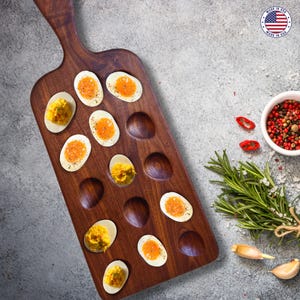 May include: A wooden serving board with egg-shaped indentations, filled with deviled eggs. The eggs are arranged on the board, alongside a small bowl of peppercorns, rosemary, garlic, and chili peppers. The board is made in the USA.