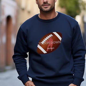 Könnte beinhalten: Ein marineblaues Sweatshirt mit einem braunen und weißen Football-Motiv. Der Football hat eine rote Silhouette eines Footballspielers im Inneren. Der Text "Touch Down Season" ist auf den Football gedruckt.