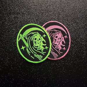 May include: Two circular embroidered patches featuring a grim reaper figure holding a scythe with small star accents. One patch has a bright neon green border and stitching, while the other has a vibrant pink border and stitching on black fabric.