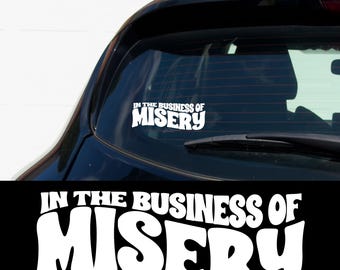 May include: White vinyl sticker with the text "In the Business of Misery" in a retro font style. The sticker is applied to the rear window of a black car.