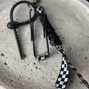May include: Black keychain with a paperclip design, featuring the letters "EMO" on small white and black beads. The keychain includes a black braided cord, a checkered black and white ribbon, and a silver clasp.