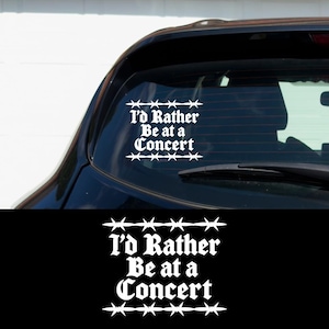 May include: White vinyl decal with the text "I'd Rather Be at a Concert" in a gothic font, framed by barbed wire designs. The decal is applied to the rear window of a black vehicle. The image also shows a black background with the same decal.