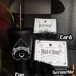 May include: A black cup with a Bridesmaid design, a black scrunchie, and two cards with Ouija board-inspired text. One card reads "Bridesmaid?" and the other reads "Maid of Honor?" The items are arranged on a reflective surface.