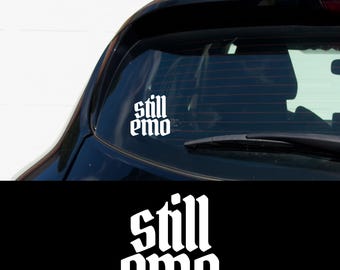 May include: A white vinyl sticker with the text "still emo" in a gothic font, applied to the rear window of a black car.