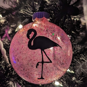May include: A pink glitter-filled Christmas ornament with a black flamingo silhouette. The ornament has a purple cap and is hanging on a black Christmas tree. The background includes other ornaments and decorative elements.