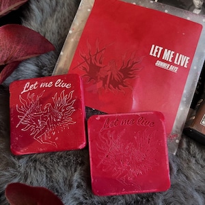 Könnte beinhalten: Rote quadratische Objekte mit dem Text "Let me live" und einer Grafik einer geflügelten Figur. Eine rote Karte mit dem Text "Let me live Summer Days" ist ebenfalls sichtbar. Die Objekte liegen auf einer grauen, pelzigen Oberfläche.