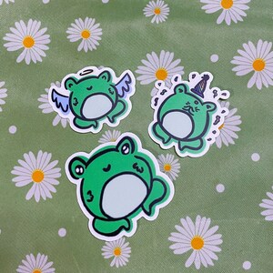 Op de afbeelding: Drie groene kikker stickers met verschillende uitdrukkingen. Eén kikker draagt een halo en vleugels, een andere draagt een feestmuts en de derde kikker glimlacht.