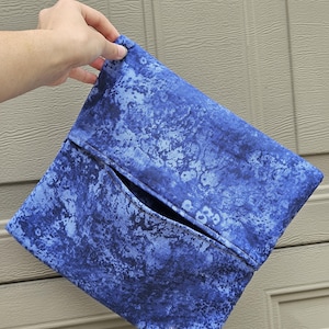 Può includere: Una pochette blu con un motivo marmorizzato. La pochette ha una tasca sul davanti ed è tenuta in mano. La tavolozza dei colori comprende sfumature di blu e bianco.