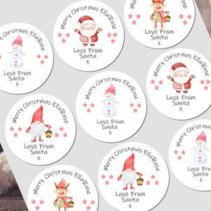 May include: A collection of white, circular Christmas gift tags with watercolor illustrations of Santa Claus, snowmen, gnomes, and reindeer. Each tag has the text "Merry Christmas Ella-Rose" and "Love From Santa X" in a handwritten style font, surrounded by pink stars.