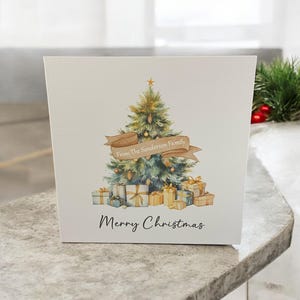 May include: A square white greeting card featuring a watercolor illustration of a decorated Christmas tree with a tan banner reading From The Sanderson Family, surrounded by gift boxes, with the text Merry Christmas printed below in black.