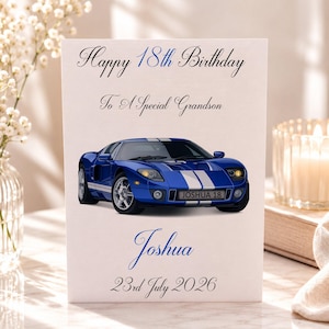 May include: A white rectangular birthday card featuring a blue sports car with white racing stripes. The text reads Happy 18th Birthday To A Special Grandson, the name Joshua, and the date 23rd July 2026. The car license plate reads JOSHUA 18.