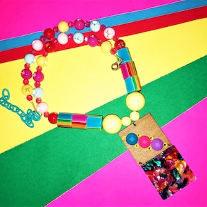 May include: A vibrant necklace with pink, yellow, and blue beads. The necklace has a rectangular pendant with a colourful, patterned design and a wooden backing. The background is a bright mix of pink, yellow, green, and blue.