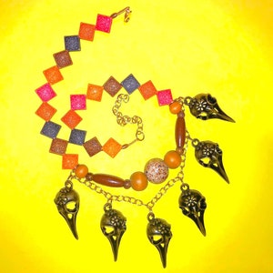 May include: A unique necklace with a spiral design. The necklace features colorful square beads in shades of pink, orange, brown, and gray. It also includes gold-colored bird skull pendants and wooden beads, all set against a bright yellow background.