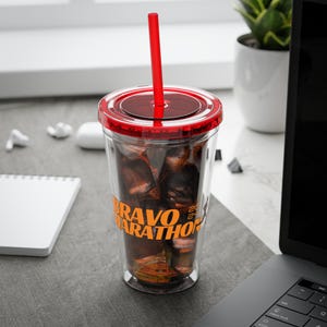 Puede incluir: Un vaso de plástico transparente con tapa roja y una pajita roja. El vaso tiene un gráfico negro y naranja que dice "BRAVO MARATHON 2023".