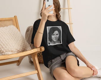 Può includere: Una t-shirt nera a maniche corte con una stampa grafica rettangolare centrata che raffigura un ritratto in bianco e nero di una persona con capelli scuri. La maglietta è indossata con pantaloncini di jeans grigi, per un look casual.