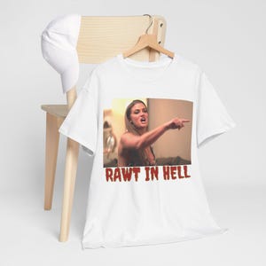 以下が含まれることがあります： 女性が指を指しているミームが特徴の白いTシャツ。赤色のテキストで「RAWT IN HELL」と書かれています。白い野球帽が背景の木製の椅子の上にあります。