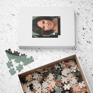 Può includere: Una scatola bianca con un puzzle da 323 pezzi all'interno. Il puzzle è di una donna con i capelli castani e una camicia bianca. La scatola ha il testo "Keep on Serving" sul lato.