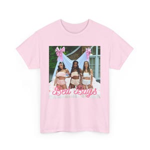 May include: A light pink t-shirt featuring a graphic of three people in lingerie on a bed, with pink bows and the text "Bed Bugs". The shirt is a soft, comfortable material.