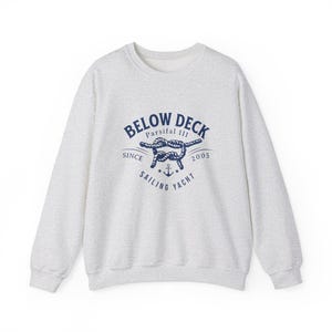 Op de afbeelding: Een grijs sweatshirt met een donkerblauw logo met de tekst "BELOW DECK Parssifal III SINCE 2005 SAILING YACHT" met een nautische knoop en anker.