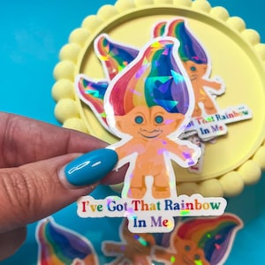 May include: A collection of stickers featuring a cartoon troll with rainbow hair and the text "I've Got That Rainbow In Me." The stickers have a holographic effect and are on a yellow tray against a blue background.