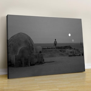 May include: A black and white canvas print depicting a desert landscape with a domed structure in the foreground. A solitary figure stands in the distance against a horizon featuring two circular suns in the sky, creating a stark, monochromatic scene.