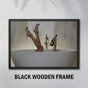 Op de afbeelding: Een ingelijste print met een persoon in een badkuip met omhooggestoken benen, zilveren hoge hakken en een champagnefles. De print zit in een zwart houten frame. De tekst onderaan luidt "BLACK WOODEN FRAME."