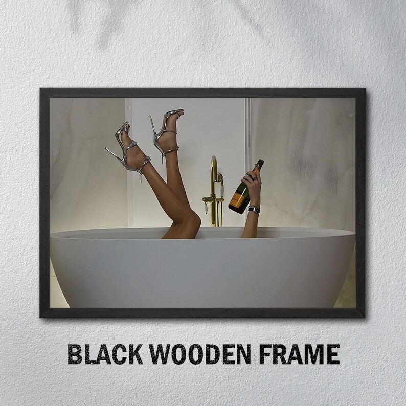 Op de afbeelding: Een ingelijste print met een persoon in een badkuip met omhooggestoken benen, zilveren hoge hakken en een champagnefles. De print zit in een zwart houten frame. De tekst onderaan luidt "BLACK WOODEN FRAME."